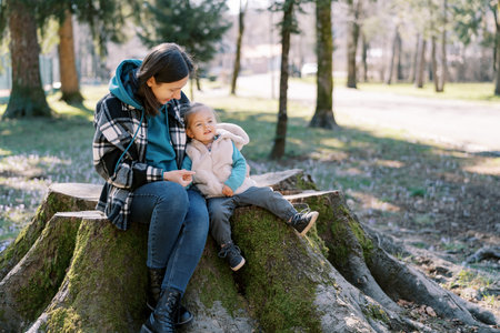 Little smiling girl sits on a huge stump leaning against her mother in the spring forest