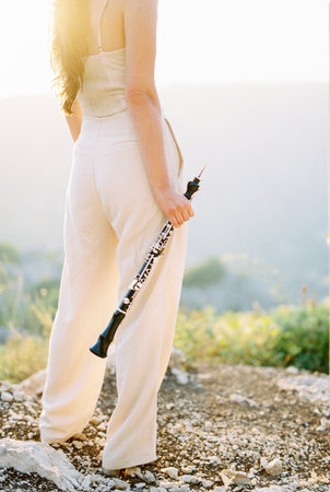 Girl with a clarinet in her hand stands on a rock and looks at the mountains. Back view. Cropped