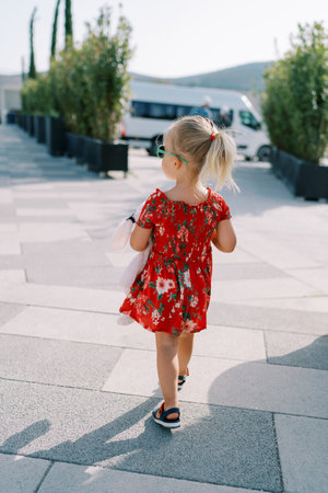 Little girl walks with a pink toy through a square with green trees in flowerpots and looks to the...