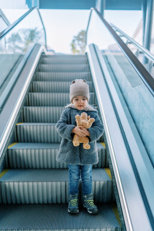 Little girl with a soft toy dog stands on an escalator and looks downの写真素材