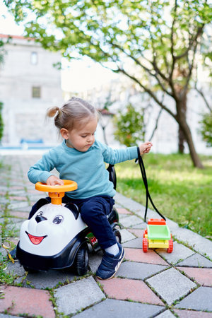 Little girl sitting on a toy car with a small toy car on a rope on a path in the gardenの写真素材