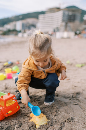 Little girl is squatting on the beach and enthusiastically digging the sand