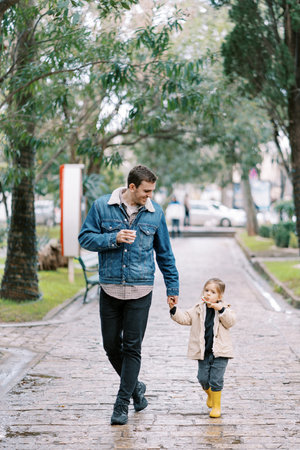 Dad with a glass of coffee walks down the street holding the hand of a little girl with a candy caneの写真素材