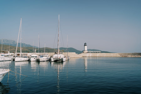 Sailing yachts and catamarans stand at the marina pier near a large breakwater with a lighthouseの写真素材