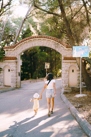 Mother with her little daughter run along the road to the arched gate in the park. Back viewの写真素材