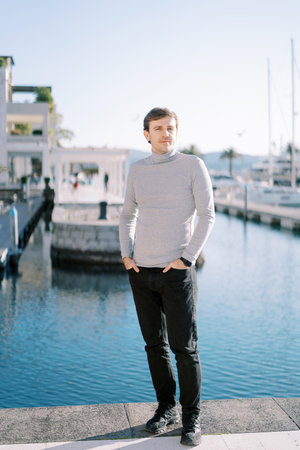 Young man with his hands in his pants pockets stands on the embankment near the moored yachts