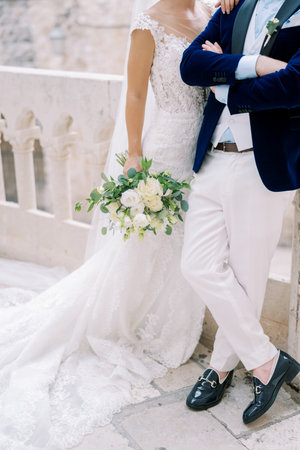 Bride with a bouquet stands with her hand on the shoulder of groom, who has his arms crossed over his chest. Cropped. Facelessの写真素材