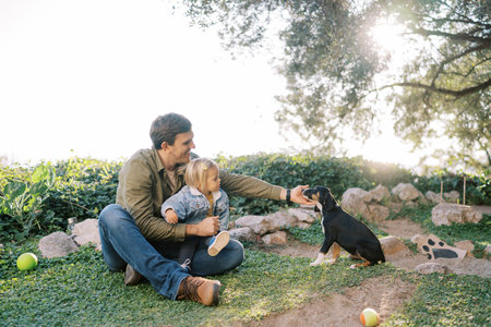 Smiling dad with a little daughter on his knees sits on green grass and pets a big puppyの写真素材
