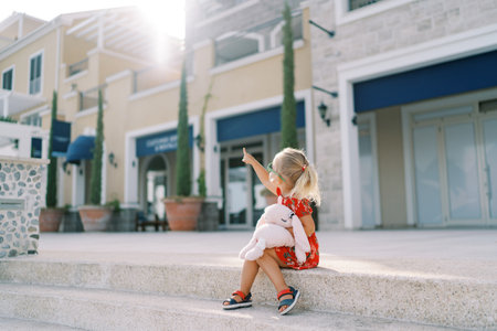 Little girl sits on the sidewalk curb and points to a house on the street. Back viewの写真素材