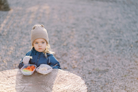 Little girl with a lunchbox stands behind a large stump and looks into the distanceの写真素材