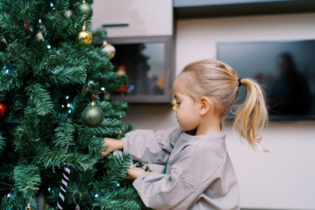 Little girl decorates the Christmas tree in the room with toys. Side viewの写真素材
