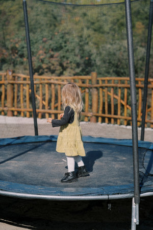 Little girl stands on a trampoline with a mesh fence in a green park. Side viewの写真素材