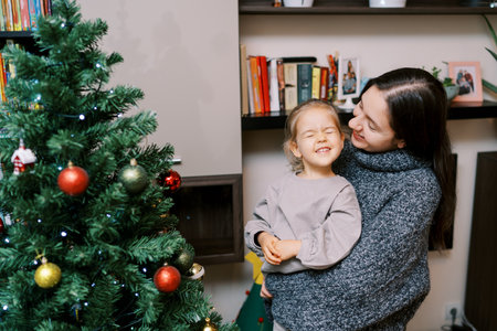 Smiling mother looks at little laughing girl in her arms while standing near decorated Christmas treeの写真素材