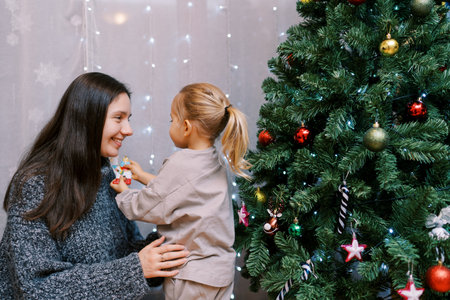 Little girl shows her mother a llama figurine while standing near a decorated Christmas tree in the roomの写真素材