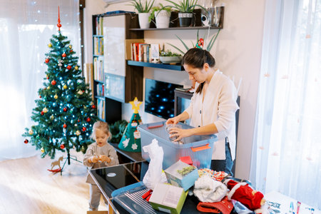 Mom and little girl sort through New Year toys on the table in a room with a decorated Christmas treeの写真素材