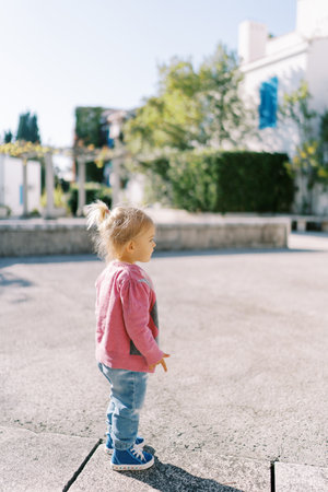 Little girl stands on the asphalt road in front of the house and looks into the distance. Side viewの写真素材