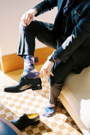 Man in suit puts on black leather shoes while sitting on bedの写真素材