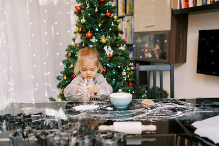 Little girl in an apron sprinkles flour on the table while sitting on a chair against the backdrop of a Christmas treeの写真素材