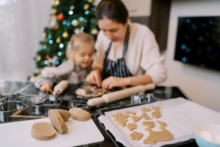 Pieces of gingerbread dough lie on a cutting board next to mom and little girl cutting out cookies with cookie cuttersの写真素材