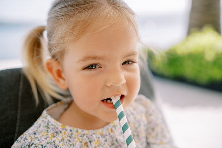 Little girl, slyly squinting, drinks through a striped straw, sitting in a chair