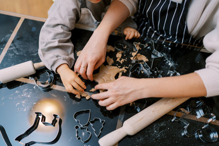 Mother and a little girl are cutting out cookies from dough with metal cutters at the table....