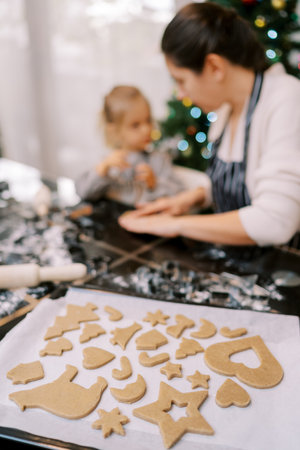 Tray of Christmas cookies stands on the table next to a mother and little girl kneading dough