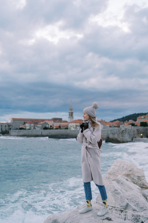 Young woman stands on a rocky shore with her coat turned up against the backdrop of the old town of...