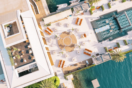 Round tables stand around a stage on a wide pier with a swimming pool by the sea. Top view. High...