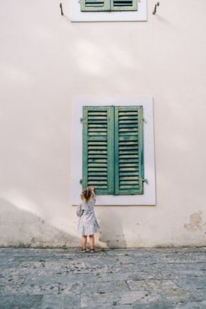 Little girl stands near an old house and touches the green wooden shutters with her hand. Back view....