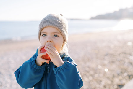 Little girl eats a big red apple holding it in her hands while standing on a sandy beach