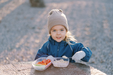 Little girl with a spoon from a lunchbox stands behind a tree stumpの写真素材