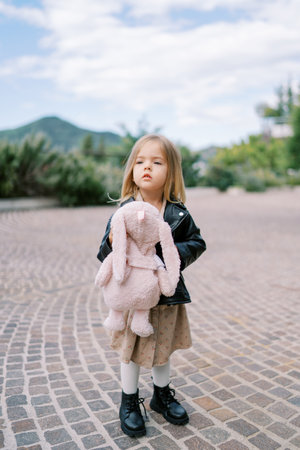 Little girl with a plush hare in her hands stands on a paved path in a green garden and looks into the distanceの写真素材