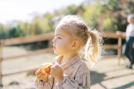 Little girl looks into the distance holding an apple in her hands while standing in the parkの写真素材