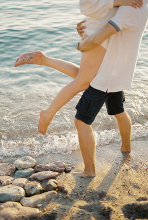 Man holds in his arms woman waving her legs while standing on the seashore. Cropped. Facelessの写真素材