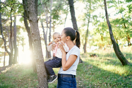 Mom kisses a little girl holding her in her arms near a tree in the parkの写真素材