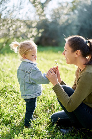 Smiling mother squatting holding hand of little girl with dandelionsの写真素材