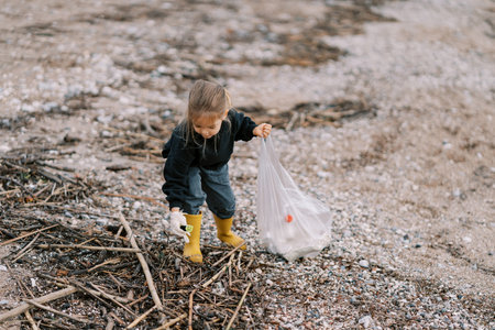Little girl bending down collects garbage on a sandy beach into a bagの写真素材