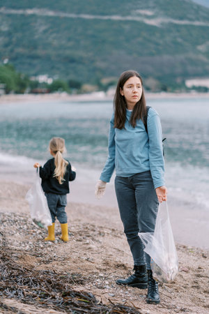Mom and little girl collect garbage in bags while standing on the beach by the seaの写真素材