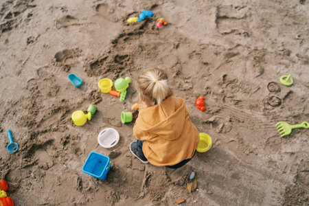 Little girl squats on the sand and plays with colorful toys. Top view