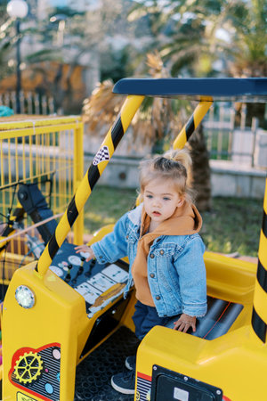 Little girl stands in the cab of a toy forklift and touches the levers of the control panel with her...