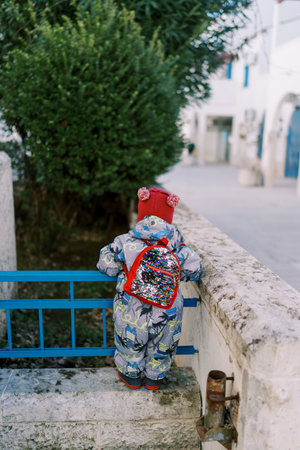 Little girl in overalls with a backpack stands on a fence near the house and looks at a tree. Back...