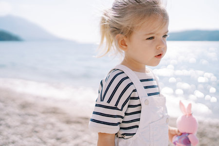 Little girl with a pink toy stands on the seashore and looks into the distanceの写真素材