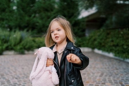 Little girl with a stuffed hare in her hands stands on a paved path in the gardenの写真素材