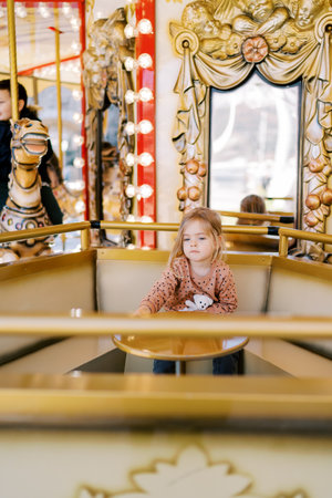 Little girl sits at a table on a carousel at a fair and looks into the distanceの写真素材