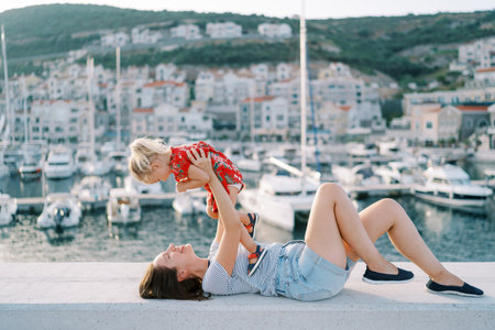 Smiling mother holds a little girl in outstretched arms while lying on the fence of the marina embankmentの写真素材