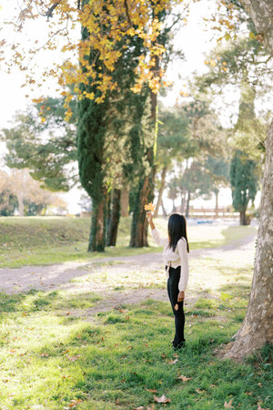 Young woman stands in an autumn park and holds a yellow leaf in her raised hand. Back viewの写真素材