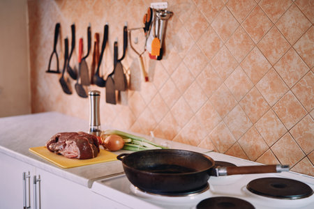 Frying pan stands on the stove in the kitchen next to raw meat and vegetables on the tableの写真素材