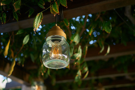 Lamp in a jar hangs on a wooden beam of a pergola entwined with green branches in the gardenの写真素材
