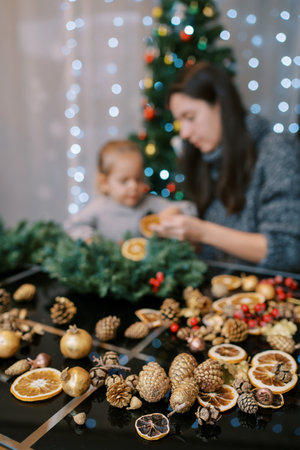 Cones and dried pieces of orange lie on the table near a mother with a little girl making a New Year wreathの写真素材