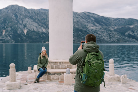 Man shoots with a smartphone a woman sitting on the step of a lighthouse on the pier. Back viewの写真素材